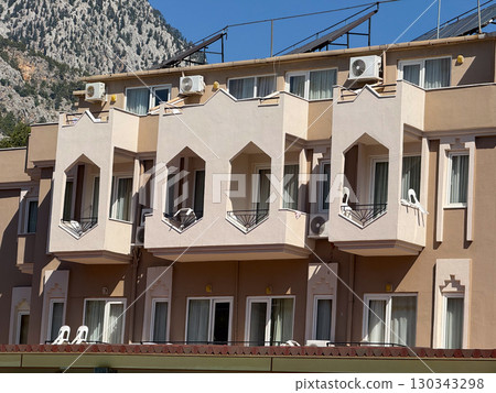 A beige building with balconies and solar panels stands before rocky mountain slopes. Architecture, urban housing and residential design blending with outdoor landscape. 130343298