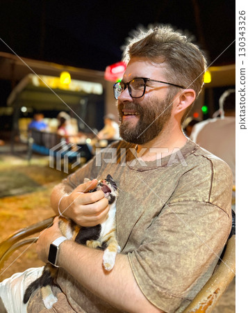 Man smiling while holding kitten in evening outdoor cafe. Human animal bond, companionship, and lifestyle with social atmosphere and emotional connection. 130343326