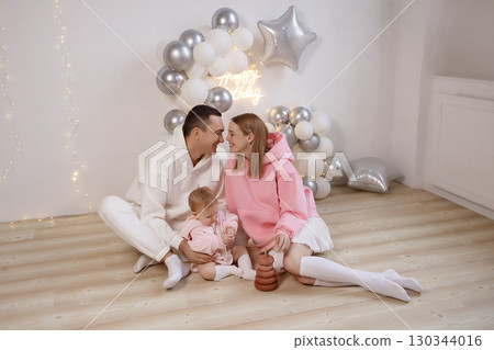 Happy young family with baby celebrating birthday on wooden floor near white wall and balloons 130344016