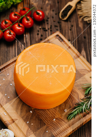 Whole round cheese wheel on wooden board with herbs and vegetables on orange background Whole round cheese wheel on wooden board with herbs and vegetables on orange background 130344372