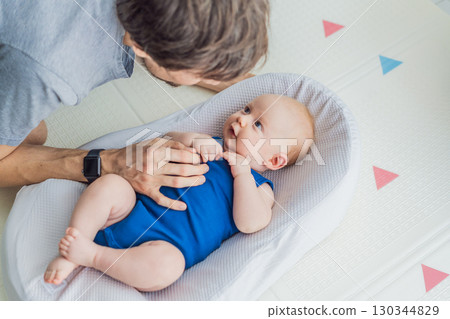 Loving father interacts with his 3-month-old baby lying in a cozy baby nest at home. Tender bonding moment, fatherhood, and early connection in a peaceful family setting 130344829