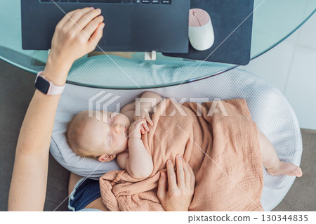 Multitasking mom works on a laptop at home while her newborn baby sleeps peacefully in a cozy baby nest on her lap. Concept of remote work, motherhood, and balancing family and career Multitasking mom works on a laptop at home while her newborn baby sleeps peacefully in a cozy baby nest on her lap. Concept of remote work, motherhood, and balancing family and career 130344835
