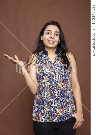 Portrait of female indian adult posing with one hand in pocket, standing against isolated background. Smiling beautiful woman in casual outfit, gesturing to side as if giving presentation in studio. 130345182
