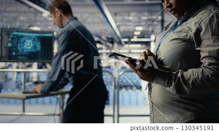 Data center programmers check AI dashboard using tablet, doing systems review. Server hub coworkers test artificial intelligence tools on device to ensure reliable performance, camera A 130345191