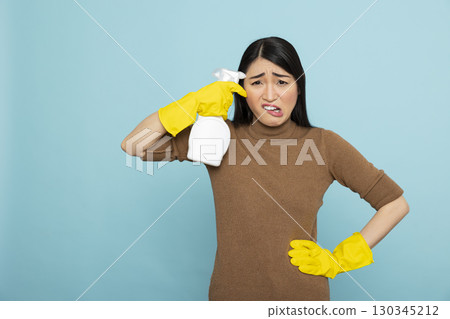 Overwhelmed asian housewife in yellow gloves holds cleaning spray bottle near forehead, expressing fatigue and frustration with household chores. Ideal for cleaning burnout concepts. 130345212