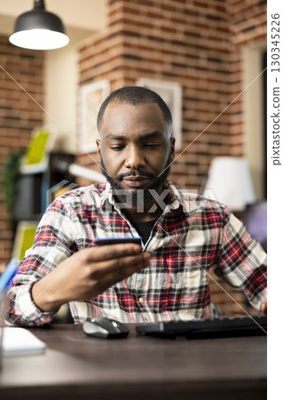 African american businessman seated at desk, reading information on credit card and completing online transactions. Black male freelancer updating his banking details on computer in brick wall room. African american businessman seated at desk, reading information on credit card and completing online transactions. Black male freelancer updating his banking details on computer in brick wall room. 130345226