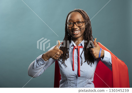 Young black woman with glasses giving two thumbs up with excitement in blue background studio. Happy african american lady in red superhero cape, expressing positivity with approval hand gestures 130345239