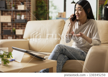 Focused businesswoman holding cellphone to ear, seated on sofa, discussing annual performance with client. Remote female manager on call with colleague, sharing updates of company financial report. Focused businesswoman holding cellphone to ear, seated on sofa, discussing annual performance with client. Remote female manager on call with colleague, sharing updates of company financial report. 130345250