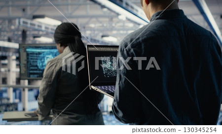 Software developer in data center checks AI dashboard using laptop during routine systems review. Server hub IT specialist tests artificial intelligence on notebook to check performance, camera B 130345254