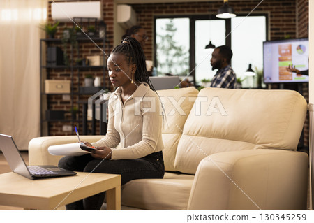 Professional woman reviewing company documents and uses laptop for data analysis in startup office. Female manager working on business project, sits at couch, using both digital and printed resources. 130345259