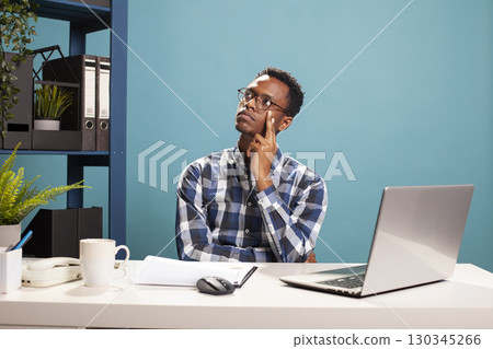 African american manager with his hand on his cheek, looking above and having deep thoughts, reflecting on office reports. Black man sitting at desk, touching his face and thinking of business ideas. 130345266