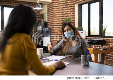 HR female representative with a pen in hand, seated at office desk conducting an interview meeting with potential job candidate. Asian businesswoman with face mask, having a discussion with employee. HR female representative with a pen in hand, seated at office desk conducting an interview meeting with potential job candidate. Asian businesswoman with face mask, having a discussion with employee. 130345292