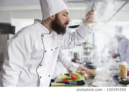 Male head chef tastes a rich aromatic sauce and testing the flavors before plating in a professional kitchen. The stovetop simmers with savory ingredients, creating a gourmet culinary experience. Male head chef tastes a rich aromatic sauce and testing the flavors before plating in a professional kitchen. The stovetop simmers with savory ingredients, creating a gourmet culinary experience. 130345300