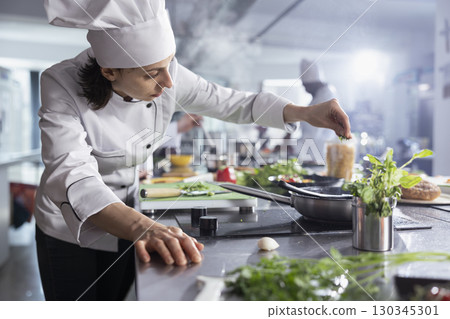 Female chef sprinkles fresh rosemary over a plated dish on the stove top, focusing on food plating and flavor. Preparing fresh ingredients, ensuring a blend of herbs in culinary workspace. 130345301