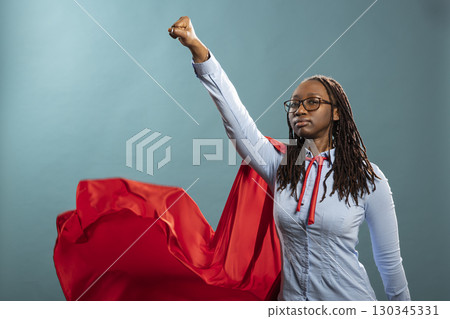 Confident woman stands in heroic stance, wearing red cape and blue shirt, exuding strength and empowerment. Black female model doing flying gesture, representing a superhero ready to save the day. 130345331