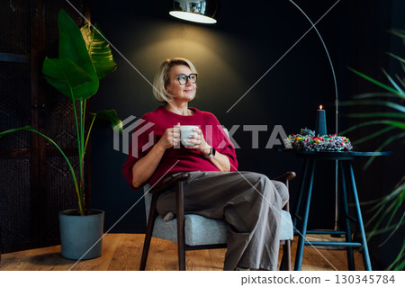 Middle aged woman relaxing with cup of hot drink in scandy style hygge interior home with winter mood decor. Lady dreaming, enjoy calm mood without stress, well being alone. Cozy fall winter holidays. Middle aged woman relaxing with cup of hot drink in scandy style hygge interior home with winter mood decor. Lady dreaming, enjoy calm mood without stress, well being alone. Cozy fall winter holidays. 130345784