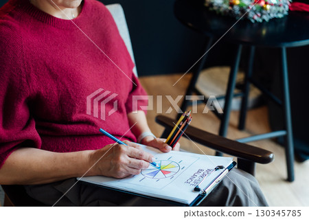 Woman drawing work-life balance wheel sitting on armchair at cozy dark home interior. Self-reflection and life planning for next new year. Personal Year review. Coaching tools. Finding Balance in Life 130345785