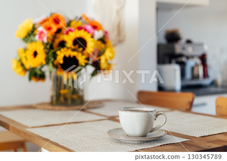 Focus on cup of coffee or tea and vase with huge sunflowers bouquet on wooden table in cozy modern interior of kitchen. Cozy Design home decor. summer mood, good morning routine. Selective focus. 130345789