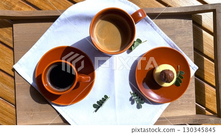 Warm morning light illuminating coffee cups and glazed chocolate cake resting on orange ceramic plate, wooden tray nearby 130345845