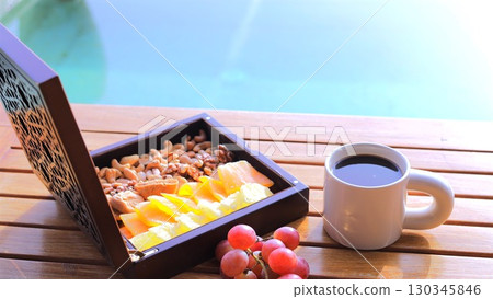 Breakfast with dried fruits and nuts in a wooden box pairs with steaming cup of coffee on a table beside turquoise pool, offering relaxing breakfast moments with another warm cup of coffee. Breakfast with dried fruits and nuts in a wooden box pairs with steaming cup of coffee on a table beside turquoise pool, offering relaxing breakfast moments with another warm cup of coffee. 130345846