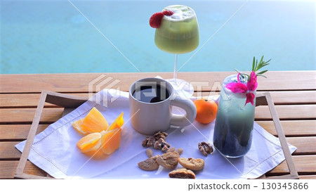 Wooden table near turquoise pool featuring refreshing breakfast spread with coffee, juice, fruits, cocktails suggesting vacation relaxation 130345866