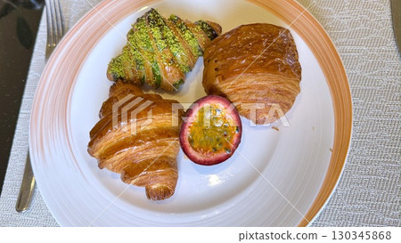 Freshly baked croissants resting on plate, croissants adorned with pistachio, alongside half passion fruit, creating inviting and delicious breakfast scene perfect for any morning. 130345868