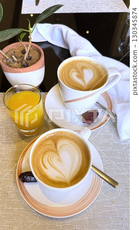 Two cappuccino cups with heart shaped latte art, paired with orange juice and dates, create cozy breakfast setting on table adorned with small potted plant 130345874