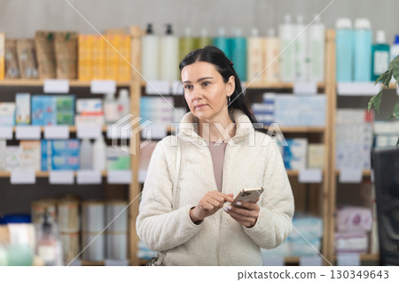 Woman is standing in a pharmacy and holding a mobile phone in her hands Woman is standing in a pharmacy and holding a mobile phone in her hands 130349643