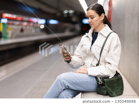 Girl with phone is sitting on metro platform station, scrolling though internet Girl with phone is sitting on metro platform station, scrolling though internet 130349898