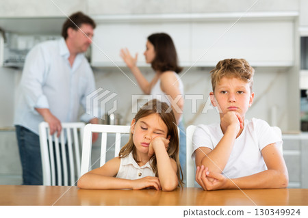 Two children sitting at the kitchen table sadly while their parents quarrelling 130349924