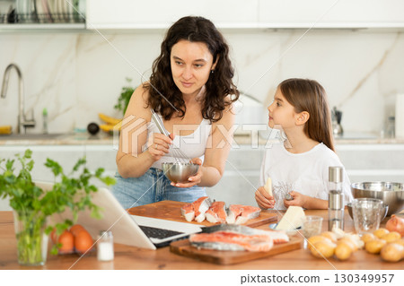 While cooking salmon fillet fish dish, woman and little daughter look at laptop 130349957
