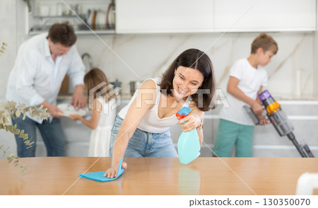 Wife cleans table with rag, husband and children clean kitchen in background Wife cleans table with rag, husband and children clean kitchen in background 130350070