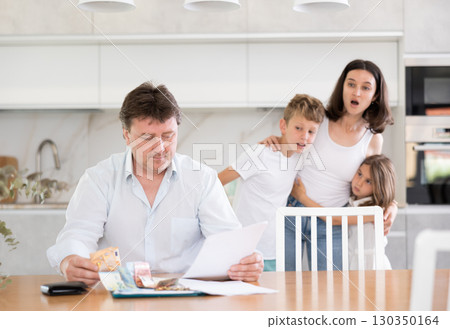 Sad father looks at bills and counts money. Upset wife with children in background Sad father looks at bills and counts money. Upset wife with children in background 130350164