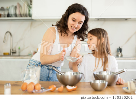 Happy mother and her daughter adding flour in metal bowl and whisking ingredients in the kitchen 130350201