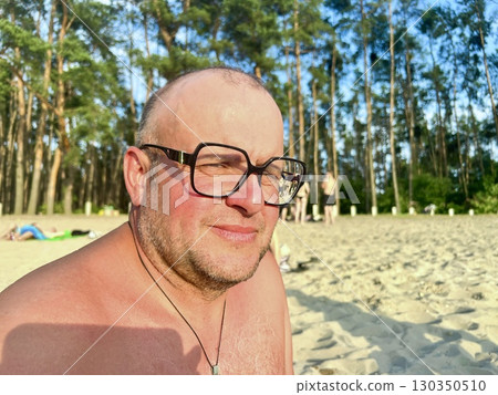 Portrait of a smiling middle-aged man in glasses relaxing on a beach by the river, enjoying summer 130350510