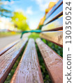 Close-up of a wooden bench in a park with a blurred background of greenery, pathway, and a sunny day 130350524