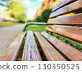 Close-up of a wooden bench in a park with a blurred background of greenery, pathway, and a sunny day 130350525