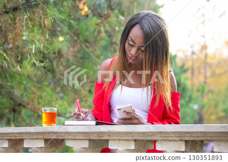 Woman in a red jacket writing notes while enjoying tea in a park during autumn Woman in a red jacket writing notes while enjoying tea in a park during autumn 130351893