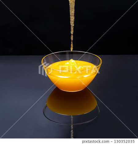 Golden syrup drizzling into a glass bowl creating a shiny reflection on a black surface 130352035