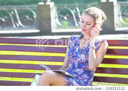 Woman sitting on a bench talking on the phone while reading a magazine in a park 130352112