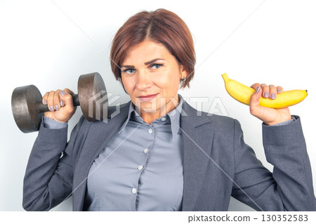 Businesswoman balances fitness and nutrition with dumbbell and banana at a light background Businesswoman balances fitness and nutrition with dumbbell and banana at a light background 130352583