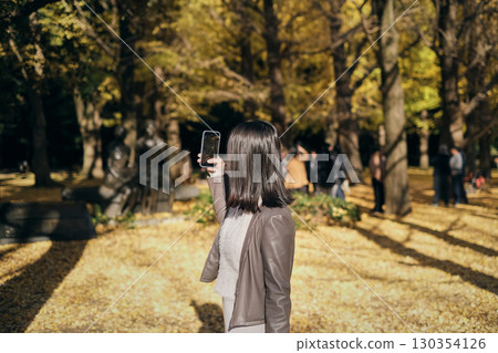 Back view of a woman taking a photo of autumn leaves with a smartphone 130354126