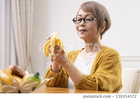 Senior woman peeling and eating a banana in the dining room 130354372