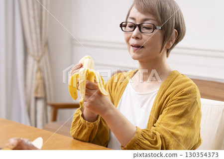 Senior woman peeling and eating a banana in the dining room 130354373