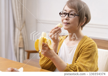 Senior woman peeling and eating a banana in the dining room 130354374