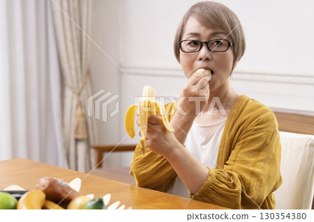 Senior woman peeling and eating a banana in the dining room Senior woman peeling and eating a banana in the dining room 130354380