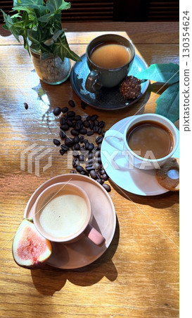 Coffee cups nestled near scattered beans, cookies, and halved fig on rustic wooden surface, evoking warm, intimate morning moment Coffee cups nestled near scattered beans, cookies, and halved fig on rustic wooden surface, evoking warm, intimate morning moment 130354624