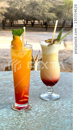 Vibrant cocktails adorned with fresh fruit and striped straws resting near grazing gazelles, blurred savanna landscape blending exotic refreshment with wildlife serenity Vibrant cocktails adorned with fresh fruit and striped straws resting near grazing gazelles, blurred savanna landscape blending exotic refreshment with wildlife serenity 130354626