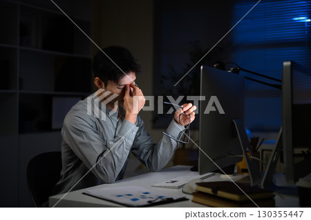 Tired businessman working late at night in office and rubbing eyes from fatigue Tired businessman working late at night in office and rubbing eyes from fatigue 130355447