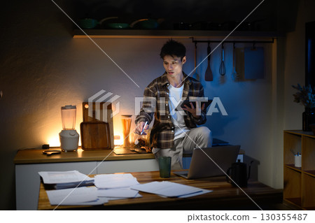 Young man working late at home, sitting on kitchen counter with digital tablet in hand Young man working late at home, sitting on kitchen counter with digital tablet in hand 130355487
